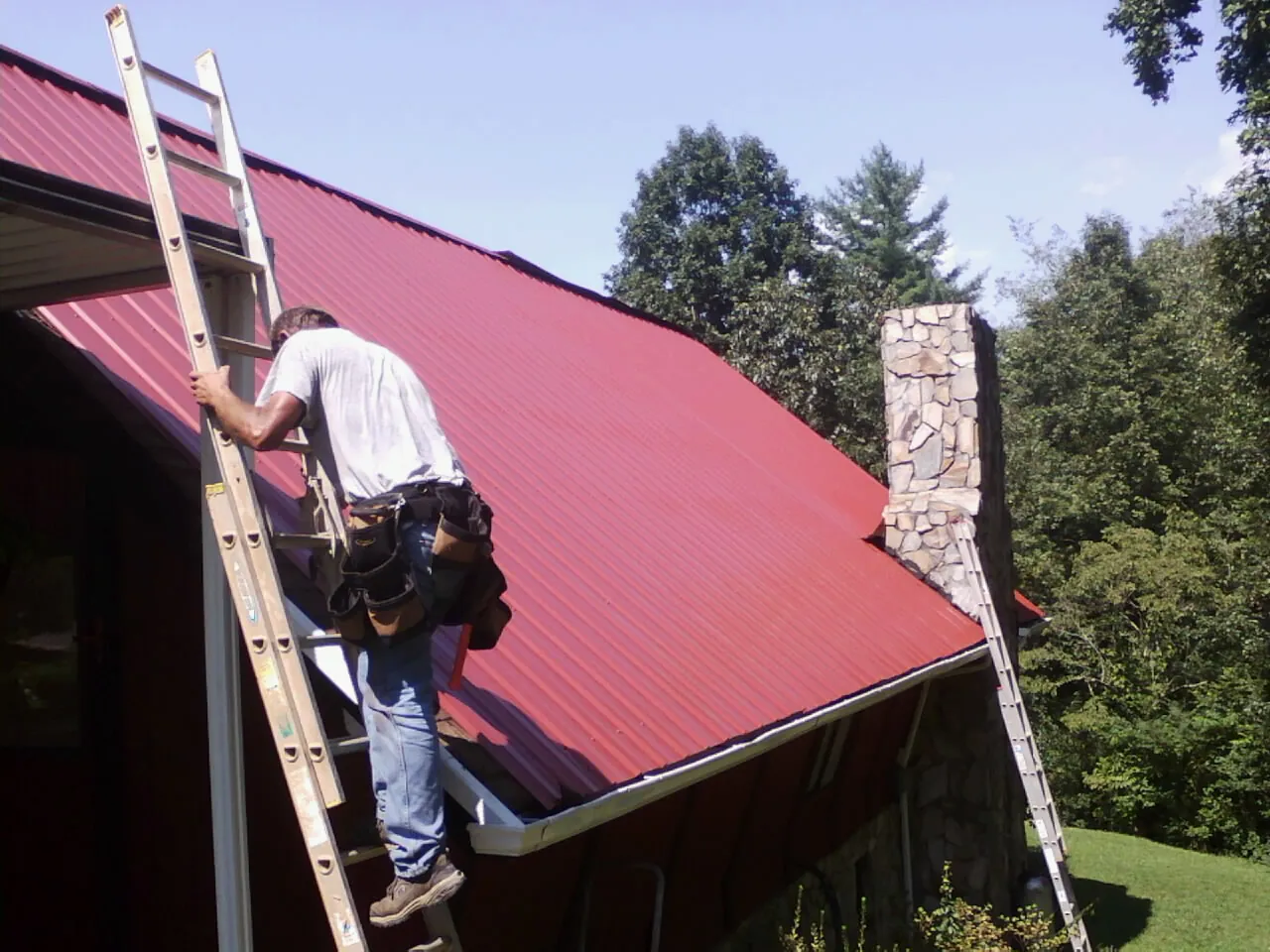Roofer on a ladder beside a red metal roof and stone chimney — Soffit & Fascia Repair by Roofing Brothers in Summer Shade